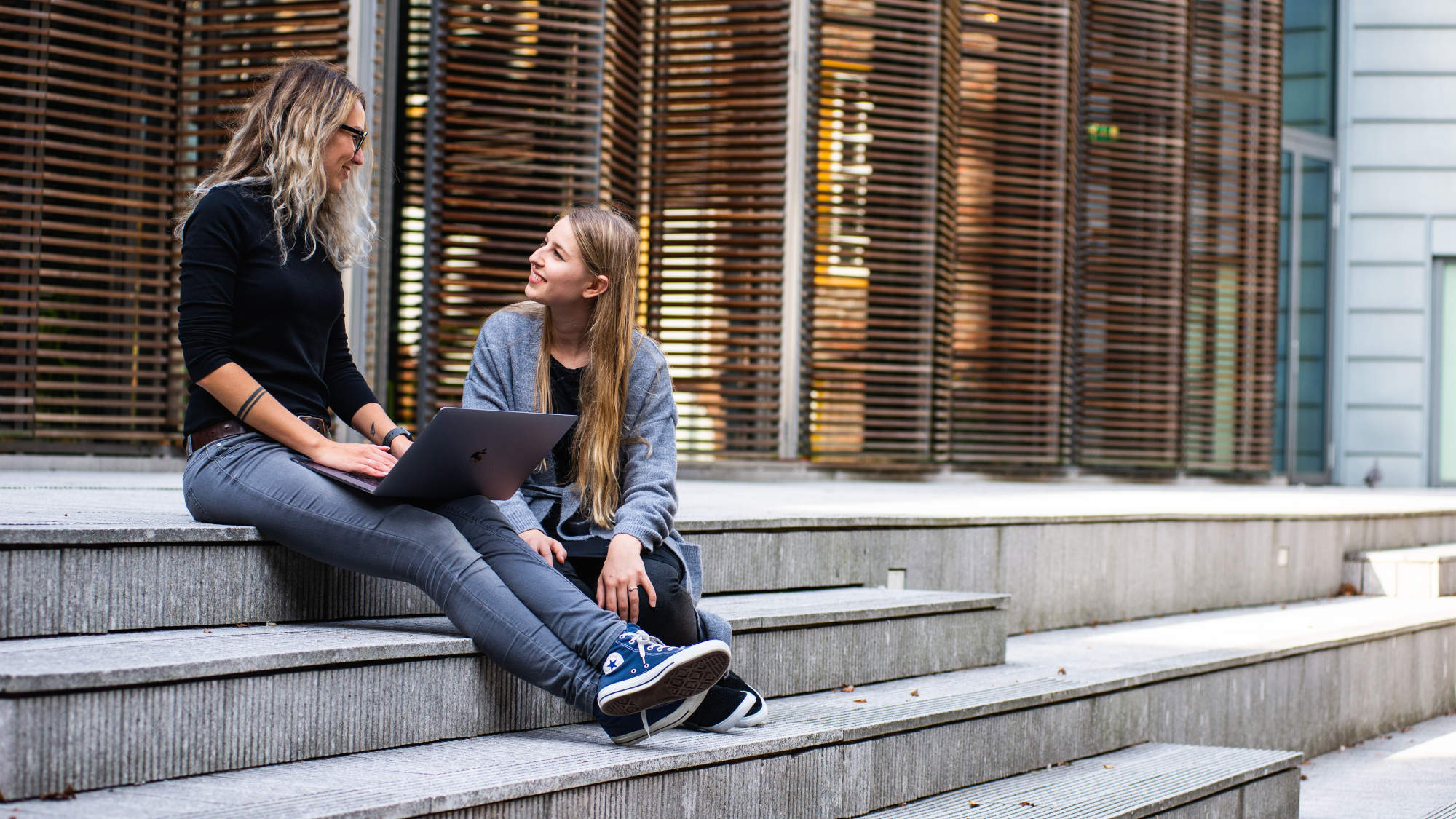 Two women sitting on stairs in front of a building and talking.