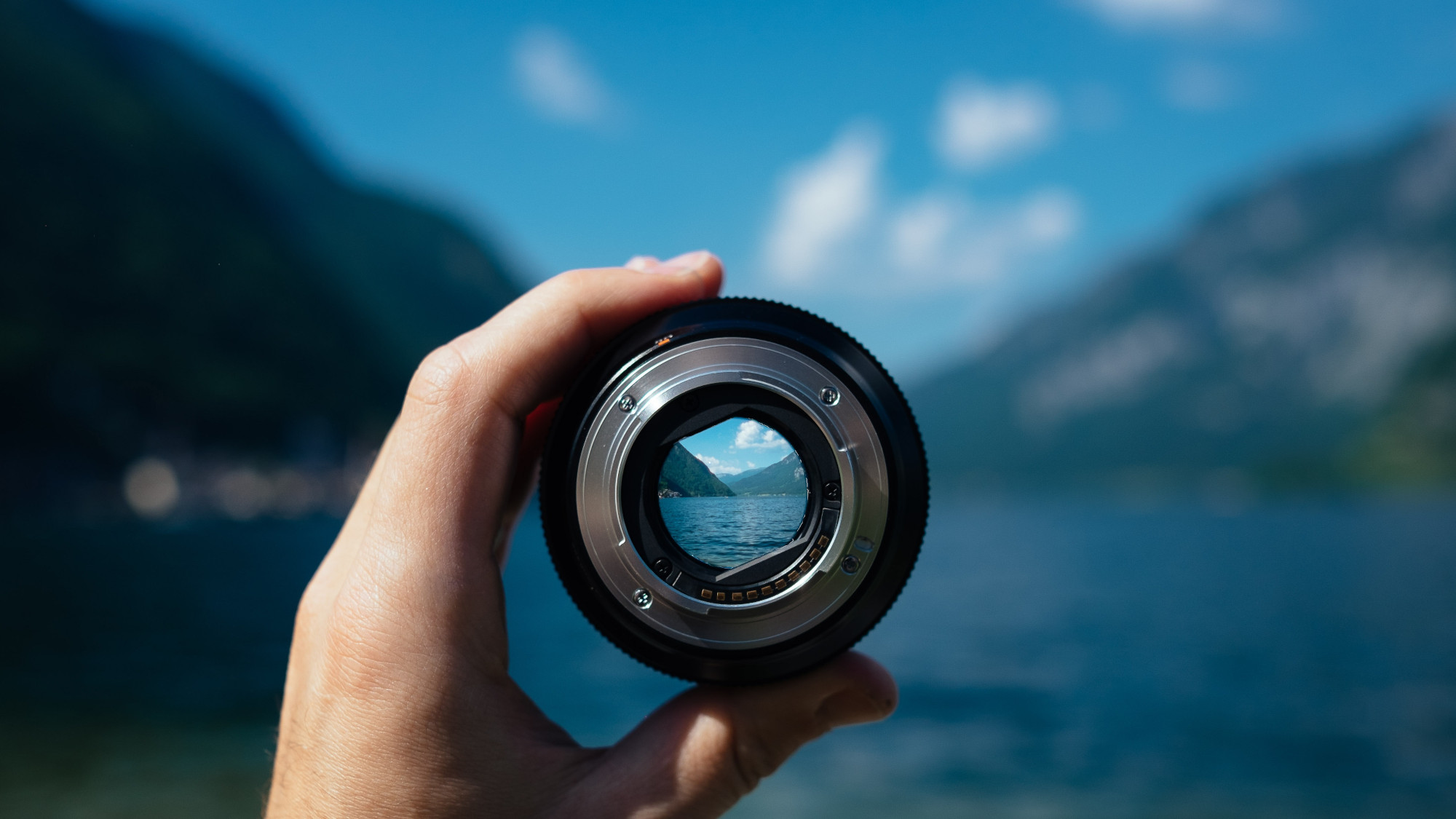 A left hand holds the lens of a photo camera in front of a blurred landscape featuring mountains, slightly rippling water and a blue slightly cloudy sky. The image inside the lens is sharp and a part of the scenery in the back can therefore be seen clearly.
