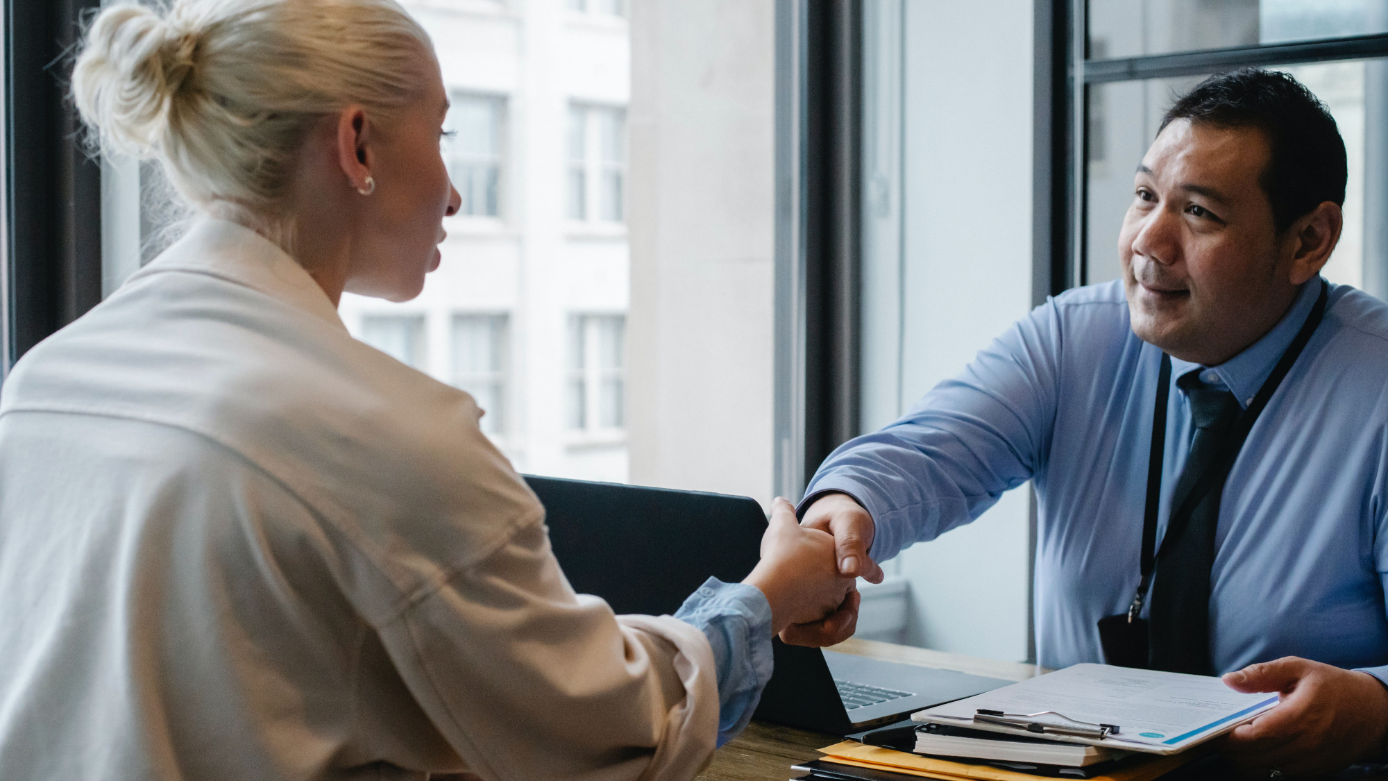 A woman and a man are sitting opposite to each other on a desk and shake hands