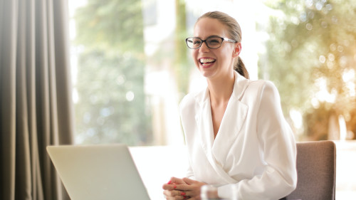 Woman with glasses dressed in a business style sits in front of a laptop smiling