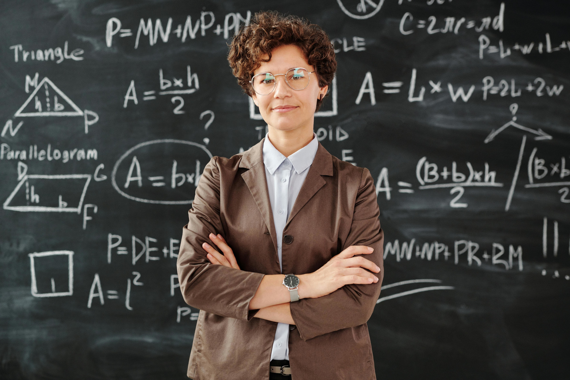 A female teacher standing in front of a blackboard which features mathematical formulars written with chalk. The teacher stands with crossed arms and is smiling.