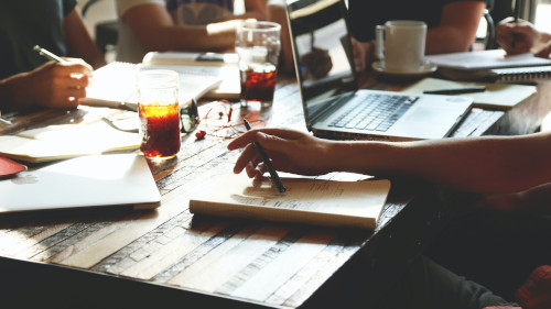 Close-up of a desk showing several notebooks and a drink in a glass. The hands of the people sitting at the table can be seen.