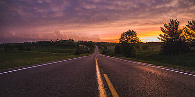 A road with grass on both sides, as well as some trees on the right side. The sun is rising and the sky is dimly lit in a purple setting.