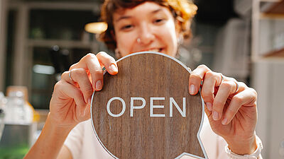 Frau die ein hölzernes rundes Schild mit dem Wort "Open" (Offen) in den Händen hält