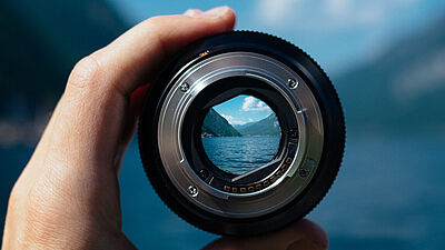 A left hand holds the lens of a photo camera in front of a blurred landscape featuring mountains, slightly rippling water and a blue slightly cloudy sky. The image inside the lens is sharp and a part of the scenery in the back can therefore be seen clearly.